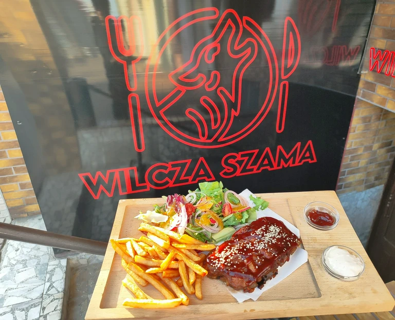 Deska z&nbsp;żeberkami w&nbsp;sosie BBQ, frytkami i&nbsp;sałatką podana przed czarną tablicą z&nbsp;czerwonym logo restauracji Wilcza Szam