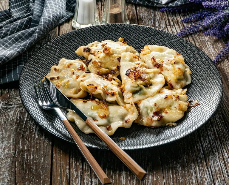 Talerz pierogów polanych podsmażoną cebulką, podanych na&nbsp;ciemnym ceramicznym talerzu z&nbsp;widelcem i&nbsp;pałeczkami na&nbsp;drewnianym stol